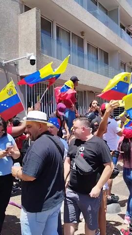 A crowd of people, many of whom are carrying Venezuelan flags, are gathered on a sunny day, presumably for a demonstration or celebration. People are dressed in casual clothes, including t-shirts, jeans, and hats. Some individuals are looking at their phones, while others are interacting with each other. The background includes buildings of varying heights, a metal fence, and some street signs.