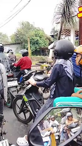 The video shows a group of motorcyclists stopped in traffic. They are all wearing helmets. The motorcyclist in the foreground is wearing a blue shirt and jeans, and a black helmet. They are looking down at their phone. In the background, there are more motorcyclists and some buildings. The traffic appears to be moving slowly. There is a red and white barrier to the left of the frame. The overall impression is one of a typical urban traffic jam.