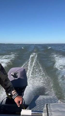 A close-up, rear-facing shot from a moving boat shows a person's arm and hand on the tiller of an outboard motor. The outboard motor, covered by a gray tarp, is at the back of the boat. The boat is creating a large wake as it moves through choppy, dark water under a clear, blue sky. The horizon is visible in the distance, with a faint line of land or trees. The overall impression is of travel on a body of water.