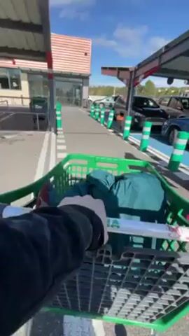 The first-person perspective video depicts someone pushing a green shopping cart, with dark green cloth inside, along a paved path lined with green and white striped bollards towards the entrance of a building. The building appears to be a supermarket or store, featuring a tiled roof, glass doors, and windows.  The path is covered and the overhead structure has red trim. Cars are parked in parking spots to the right of the path. The sky is blue with some clouds. The person pushing the cart is wearing a dark jacket.