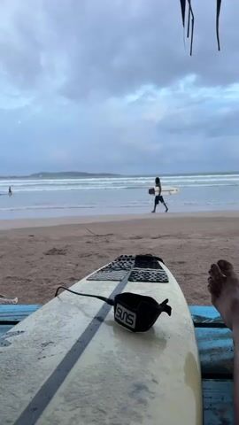 A close-up shot of a surfboard with a leash attached faces the ocean at Weligama, Sri Lanka. The board lies on a rustic, blue wooden surface, possibly a bench or table, with a black object, perhaps a helmet, to its left. In the background, a person walks along the wet sand, carrying another surfboard. The ocean is calm with small waves, and a few other figures can be seen in the water. The sky is overcast with dramatic, grey clouds, suggesting a moody, late afternoon or early morning atmosphere. The overall scene evokes a sense of tranquility and the anticipation of surfing, despite the cloudy weather. The word "SUNS" is visible on the leash of the surfboard in the foreground.