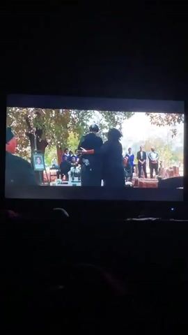 The video shows a dark room where a television screen displays a scene of a funeral taking place outdoors. Several people are gathered, and the focus is on two figures in the foreground who appear to be walking towards the ceremony. A portrait stands to the left of the frame, likely of the deceased. Behind the central figures, individuals are seated and standing, dressed formally. The background features trees and an open area. The lighting is bright in the scene on the screen, contrasting with the darkness of the room where the video is being recorded. The television emits a soft blue light from its base. In the foreground of the recording room, parts of a person and furniture are visible, adding to the ambiance of someone watching television in a dimly lit space.