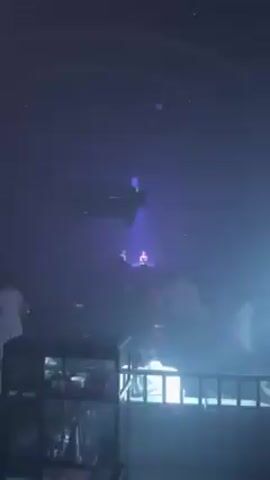 The scene is a dimly lit nightclub or event space filled with people. A stage is visible with two figures illuminated by a bright white spotlight, suggesting they are performers. An overhead structure, possibly lighting equipment or a sound system, hangs from the ceiling. The overall atmosphere appears energetic with purple and pink lighting adding to the ambiance. The crowd is moving and seems to be enjoying the performance.