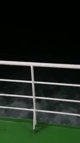 The video shows a nighttime scene on a boat, with a view from behind a white metal railing. The deck of the boat is green. The water is dark and agitated, with white foam visible. The sky is black.