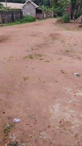 The video shows a dirt road in a rural area. The road is reddish-brown and has patches of grass and litter. In the background, there are buildings, trees, and a water tower with solar panels. The sky is cloudy.
