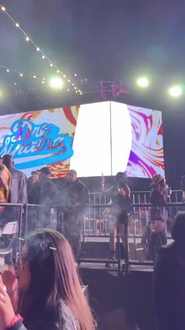 The scene depicts a music concert or festival at night. A large LED screen in the background displays the name "Dre Sinatra" in a stylized font over a swirling, psychedelic pattern. Below the screen, a crowd of people stands, some appearing to be on a raised platform or VIP section behind metal barriers. Several individuals in the crowd are actively engaging with the music, raising their hands and dancing. The lighting includes overhead string lights and stage lights, creating a vibrant and energetic atmosphere. The people in the foreground are dressed in various casual attires, capturing the youthful and lively energy of the event.