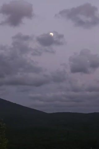 The video shows a time-lapse of the moon in a cloudy sky over a dark, forested mountain range. The moon is visible in the upper center of the frame, surrounded by various shapes and sizes of gray clouds. The foreground is dominated by the silhouette of the mountain, which appears to be covered in dense foliage. The overall mood is serene and slightly dramatic due to the twilight lighting and the overcast sky.