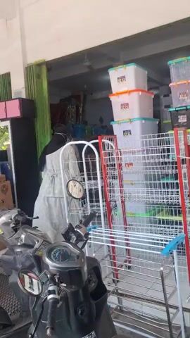 The video shows the exterior of a store with a motorcycle parked in the foreground. Inside, various items are displayed, including stacked plastic storage bins of different colors, wire shelving units, and a tall, black and white cabinet topped with a large red pot. A folding green door is partially open, revealing more items inside the store. A security camera is visible mounted on the wall. The overall scene appears to be an outdoor market or retail space selling household goods.