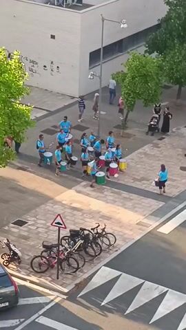 A group of people, mostly young adults, are playing drums on a paved street in what appears to be a city. The drummers are wearing blue shirts and black shorts and are arranged in a semi-circle formation, facing a woman who is standing to the right and playing a drum. The drummers are playing a variety of drums, including large bass drums and smaller snare drums. Some of the drums are brightly colored, with green, pink, and yellow drums visible. In the background, there are buildings, trees, and a few spectators watching the performance. There are also bicycles parked on the side of the street and a car parked at the bottom of the frame. The sun is shining, casting long shadows across the street. The overall scene appears to be a public performance or parade.