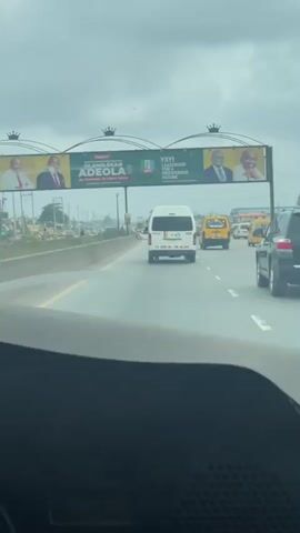 The video shows a highway scene. In the foreground, a dashboard is visible, suggesting the footage is filmed from inside a vehicle. The road ahead is lined with multiple lanes of traffic. Several vehicles are in motion, including a white minivan, a yellow taxi, and a black SUV. In the background, a large billboard is prominently displayed over the highway. The billboard features portraits of two men, accompanied by text that appears to be political in nature, mentioning "YAYI LEADERSHIP FOR A PROSPEROUS FUTURE" and "OLAMILEKAN ADEOLA." The sky is overcast, indicating a cloudy day. The environment appears to be urban or suburban, with some structures visible in the distance.