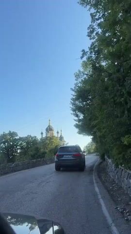 The video shows a view from a car traveling on a road with a stone wall on the left and trees on the right. A dark colored SUV is ahead of the camera vehicle, also traveling on the road. In the distance, to the left of the road, a church with multiple domes and crosses is visible, partially obscured by trees. The sky is clear and blue.