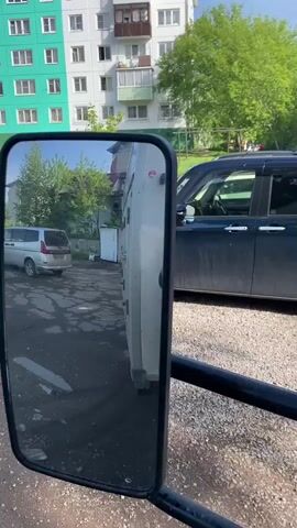 The video offers a close-up view through a vehicle's rectangular side mirror, with the mirror itself occupying the left side of the frame. Reflected in the mirror, a white truck or large van is prominently featured, parked or moving slowly. To its left in the reflection, a silver van is observed driving away on a paved road. Beyond these vehicles, a multi-story apartment building with white and green sections is visible, along with lush green trees under a bright, sunny sky. On the right side of the main frame, outside the mirror's reflection, a dark blue car is parked on a gravel or dirt lot, with other vehicles parked further in the background. The scene depicts a daytime setting in what appears to be a residential area. No legible text is visible in the video.