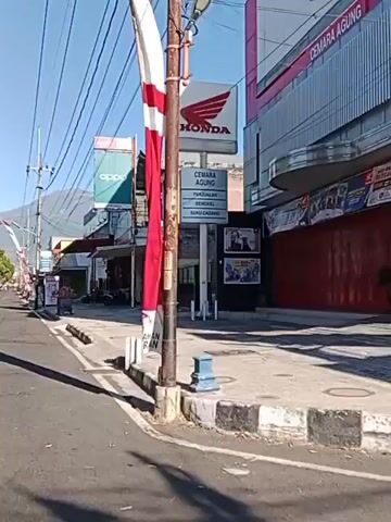 The video pans along a street in what appears to be a commercial area. The street is paved with asphalt and has a curb on the right side. There are several buildings lining the street, most of which appear to be shops. Signs for various businesses are visible, including Honda, Oppo, and Fastron Auto Service. Several motorcycles are parked along the sidewalk. A red and white flag is prominently displayed on a pole in the foreground on the right. The sky is blue and clear, and there are mountains in the background. The overall impression is that of a sunny day in a busy urban or semi-urban environment.