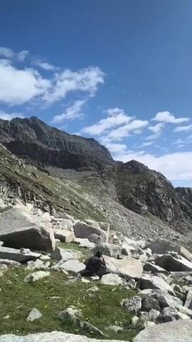 A vertical video shows a person sitting on a boulder in a rocky, mountainous landscape under a blue sky with scattered clouds. The person, dressed in dark clothing, is facing away from the camera. The environment consists of large rocks, sparse green vegetation, and steep, rocky mountain slopes in the background. As the video progresses, the camera pans slightly to the right, revealing more of the mountain range and a valley filled with clouds. The sun is visible in the upper right corner, casting light on the scene.
