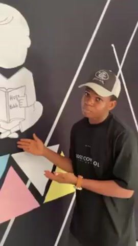 A young man is standing in front of a black wall with a drawing of a person reading a book labeled "Holy Bible." The wall also features colorful triangles and white lines. He is wearing a black t-shirt with the words "LOST CONTROL," a two-toned baseball cap, and a wristwatch. He gestures with his hands, seemingly presenting the drawing on the wall, with a slightly quizzical expression. The background suggests an indoor location with a busy atmosphere, possibly a public space.