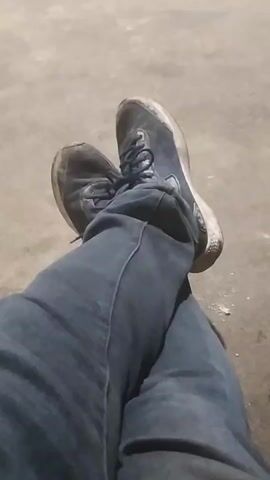 The video shows a close-up view of a person's legs, wearing blue jeans and dark sneakers. The person's legs are crossed, with one foot resting on top of the other. The background is a neutral, textured surface that appears to be concrete or asphalt. The lighting is dim, suggesting it might be evening or indoors. The camera is positioned above and slightly to the side of the person's legs, providing a clear view of their attire and posture. There is a small white object on the ground near the person's feet. The video is brief and static, with no discernible movement or action beyond the static presentation of the legs and feet.