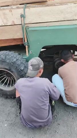 Three individuals are crouched down next to the rear axle of a green pickup truck. The truck bed is loaded with several light-colored wooden planks secured by a rope and a wooden pole. The ground appears to be a dirt or gravel surface. The individuals are dressed casually: one in a gray t-shirt and baseball cap, another in a tan t-shirt and light blue pants, and the third in a black t-shirt and dark shorts. The person in the tan shirt and blue pants is looking towards the truck's axle, while the person in the black t-shirt is reaching towards the axle with a tool, possibly a wrench. The person in the gray shirt is looking down at their hands. They appear to be working on the truck's rear wheel or braking mechanism.