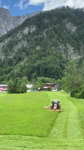 A red and white tractor with a mowing attachment is cutting a field of green grass. The tractor is moving from left to right across the frame. The grass is being cut into rows, creating a patterned effect. In the background, there are several houses and trees. Beyond the houses and trees, there is a large, steep, rocky mountain covered in green vegetation. The sky is blue with some white clouds. The sun is shining. The video is shot from a high angle, looking down on the scene.