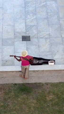 The video shows an overhead shot of a person playing an acoustic guitar. The person is standing on a concrete ledge with a grassy area below. They are wearing a wide-brimmed hat, a pink top, and striped pants. To their right, there is an open guitar case with a white container inside. The person is strumming the guitar and appears to be looking down at the instrument. The background is a wall made of large, light-colored tiles. The lighting suggests it is daytime.