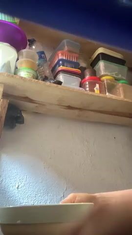 A person washes a bowl in a kitchen. The person’s hands hold a tan-colored bowl. They vigorously swirl water inside the bowl, then pour the water out. Behind them, an unevenly stacked shelf displays containers of various sizes, colors, and materials. The shelf is made of light-colored wood. The wall behind the shelf is light-colored.