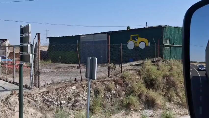 The video shows a roadside view of a long, green corrugated metal building with a yellow drawing of a roller machine on its side. The building is surrounded by a chain-link fence and appears to be an industrial or storage facility. The area around the building is dry and arid, with sparse vegetation. The video is shot from a moving vehicle, as indicated by the reflection of the vehicle's side mirror in the corner of the frame and the blur of the road ahead.