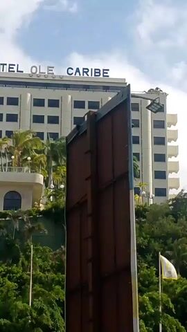 A video shows the facade of a hotel with the sign "HOTEL OLE CARIBE" on its roof. The hotel is white with dark windows. In front of the hotel, there is a tall brown structure, possibly a billboard or a wall, obscuring part of the view. Green trees and palm trees are visible on a slope leading up to the hotel. The sky is cloudy.