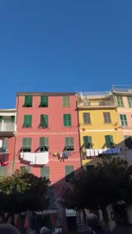 The video pans across a harbor scene in a European village. It begins with a view of colorful buildings in the background, featuring a red building with green window shutters and a yellow building with brown shutters. Laundry is hanging out to dry on the red building. The camera pans to reveal a yellow church steeple with a clock, set against a green hillside. Then, the camera shows a view of the harbor, with a cement walkway, trash receptacles, and a lamp post in the foreground. Further away, a breakwater is visible, with a number of people standing on it. Various boats are docked in the harbor. The backdrop of the harbor includes a mountainous landscape. The sky is clear and blue.