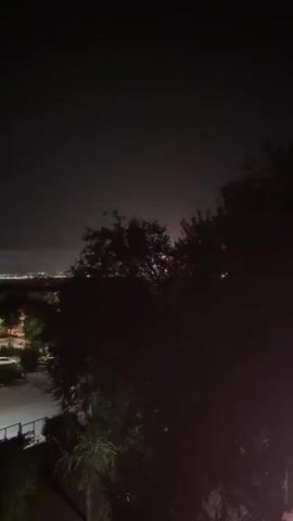 The video shows a nighttime scene with trees in the foreground obscuring a distant cityscape. There are flashes of light in the distance, possibly fireworks or lightning. The sky is dark with some clouds. The overall impression is of a quiet, somewhat eerie night.