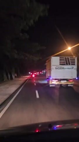 The video shows a nighttime view from inside a vehicle on a paved road. The vehicle appears to be moving, as indicated by the motion blur. On the right side of the road, a large white truck with yellow trim is visible, its taillights and other lights illuminated. The truck has writing on its side that is partially legible. Ahead on the road, other vehicles with red and white lights are visible. The surroundings are dark, with trees lining the left side of the road and streetlights providing some illumination. The road has white lane markings. The camera is positioned looking forward and slightly to the right. The overall impression is a drive on a road at night, possibly in an urban or semi-urban area.