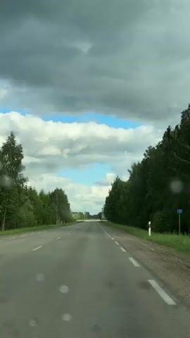 The video shows a view from inside a car driving down a straight asphalt road. The road is flanked on both sides by dense green trees. The sky above is predominantly covered in grey and white clouds, with patches of blue sky visible between them. The road markings are white, indicating lanes and the roadside edge. The windshield has a few water droplets on it, suggesting recent rain. There are no other vehicles or people visible in the video.