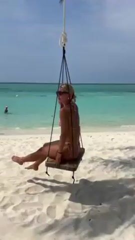 A woman in a pink bikini sits on a wooden swing hanging from a rope on a sandy beach. She wears sunglasses and has blonde hair pulled back.  Behind her, the ocean is turquoise blue, with a person visible swimming in the distance. Shadows from the tree providing the swing are visible on the sand. The sky is mostly clear with a few clouds. She touches her face and looks around.