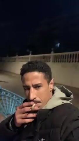 A man with short, curly black hair stands outside and holds a lit cigarette, looking at the camera. He is wearing a black puffy coat with a light gray hoodie underneath. The camera pans up and reveals a wrought iron bench and a second man sitting on the bench, wearing a gray shirt and jeans. The background shows a street at night, illuminated by street lights. The first man exhales smoke into the camera.