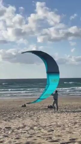 Here is a description of the video.
The video shows a man standing on a sandy beach holding a large blue and black kite. The kite is arched and partially inflated. The man appears to be preparing to kitesurf.  The ocean is visible in the background with waves rolling towards the shore. The sky is blue with scattered white clouds. The kite is marked with the word "AIRUSH."  The man walks forward and the kite rises into the air. The camera follows the kite as it flies higher, obscuring the man.