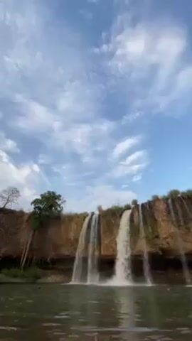 The video shows a waterfall with multiple streams of water cascading over a rocky cliff into a body of water. The sky is blue with scattered white clouds. There is some sparse vegetation on top of the cliff and a few trees to the left of the falls. The water in the pool below appears somewhat murky. The camera is stationary and captures a wide view of the scene.