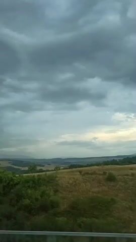 The video shows a landscape with rolling hills covered in dry grass and green bushes. The sky is overcast with dark clouds. The perspective is from a moving vehicle, likely a car, as evidenced by the blurred foreground and the slight movement of the scenery. No people or specific objects are clearly identifiable beyond the natural landscape. The scene suggests a rural environment under a cloudy sky, possibly before or during a storm.