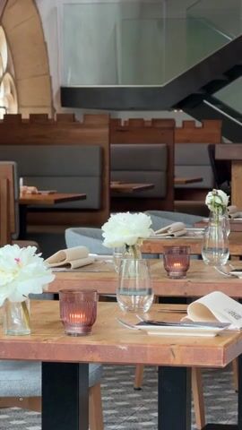 The interior of a restaurant is shown. Several wooden tables are set with glassware, white flowers in vases, napkins, silverware, and small candle holders. Behind the tables are booths with light gray upholstery. In the background, an archway and a modern staircase are visible. The setting suggests a quiet, upscale dining environment.