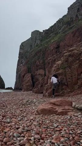 A person with dark, curly hair, wearing a white shirt and grey pants, climbs a cliffside composed of reddish-brown rock. The cliff descends to a beach covered with red and grey rounded rocks. To the left, a rocky formation stands in the water. The sky is overcast.
