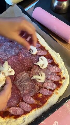 A close-up overhead shot shows hands preparing a pizza. The pizza base is spread with red sauce and topped with slices of salami and mushrooms. The hands are carefully arranging slices of mushroom onto the pizza. In the background, a pink rolling pin and a metal pot are visible. The pizza is on a baking sheet lined with parchment paper.