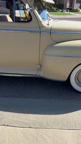 The video shows the side of a tan 1940s sedan parked on a street. The camera pans across the car, focusing on the doors, windows, and the front fender with a whitewall tire. A person's hand with tattoos is holding a camera, suggesting they are filming the car. The environment appears to be outdoors on a sunny day with a clear blue sky.