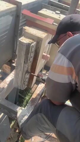 A person wearing a black baseball cap, glasses, a grey t-shirt with orange and white stripes, and grey pants is working on a construction site. The person is sitting on a metal structure and is using a drill to cut into a concrete block. The concrete block is supported by metal beams and appears to be part of a larger construction project. In the background, there are more metal structures, possibly part of industrial machinery or scaffolding, and some greenery. The scene is outdoors, and the lighting suggests it is daytime, possibly a sunny day. The person's focus is on the task at hand. The setting appears to be an industrial or construction area in Pivdennoukrainsk, Ukraine. There is no visible text in the media.