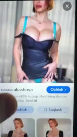 A phone screen is visible in the video, displaying a search results page in Uzbek for "casca akashova." The top result is an image of a woman in a black corset-style top, with a glimpse of turquoise underwear visible. Below this are additional images of the same woman, primarily in low-cut dresses. A finger touches the screen, indicating interaction with the search results. The lighting appears to be indoors.