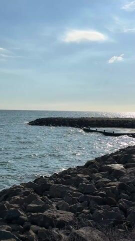 The camera pans left to right over a rocky shoreline, with water lapping against the rocks and a small concrete platform. The sky is blue with a few white clouds, and the sun is reflecting off the water.  In the distance is a hazy view of buildings.