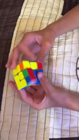 A close-up shot shows a person's hands manipulating a three-by-three Rubik's Cube. The cube is blurred in motion in the beginning of the clip, then comes into focus, showing its various colored sides. The hands are seen rotating sections of the cube. The background is a textured fabric, possibly a blanket or comforter, with vertical stripes of varying shades of brown and cream.
