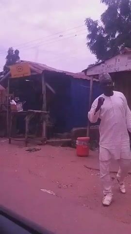 The video shows a street scene with several market stalls. A person is riding a motorcycle carrying a large bag. There are also other people standing around the stalls, some of whom appear to be customers or vendors. The stalls have tin roofs and are decorated with yellow signs bearing the MTN logo, indicating telecommunications services are offered. The ground is reddish-brown, suggesting a dirt or unpaved road. The sky is overcast with a purplish hue. The overall atmosphere appears to be that of a bustling, open-air market.