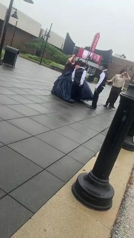 A group of people in formal attire are gathered on a wet tiled plaza in front of a mall entrance. A person wearing a large dark blue formal gown is in the center of the group. Other members of the group are wearing suits and vests. Behind them, the Woodfield Mall sign is visible on the building. The sky is overcast.