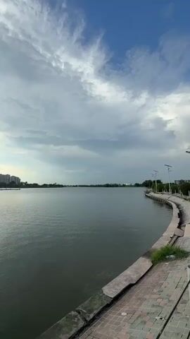 A vertical video shows a body of water with a cityscape in the background under a cloudy sky. The water is dark and appears calm, with ripples visible on the surface. A paved walkway with lights runs along the right side of the water, curving out of view. The cityscape consists of tall, modern buildings lined up along the far bank of the water. The sky is partly cloudy with some patches of blue visible. The sun is obscured by clouds, but its light casts a bright glow in the distance. The overall impression is one of a tranquil urban waterfront.