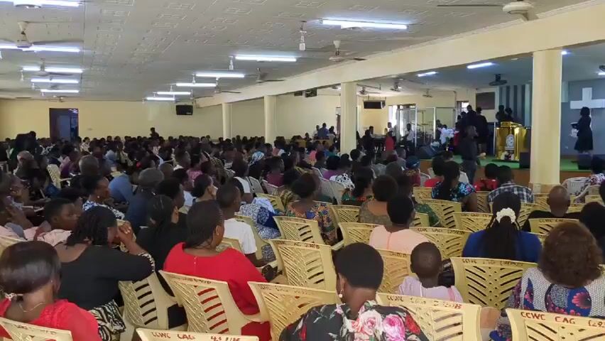 Alright, here is a concise description of the video:

The video shows a large congregation of people seated in what appears to be a church or large meeting hall. The room is well-lit with fluorescent lights and ceiling fans. Most attendees are sitting on yellow plastic chairs, facing towards a stage area at the front of the room. On the stage, there is a group of people, some dressed in formal attire, near a podium. There's a cross visible in the background. The people in the audience are diverse in age and are wearing a variety of clothing, many with colorful patterns. A man in a bright floral shirt stands out near the camera. A camera person is present, indicating the event is likely being recorded.