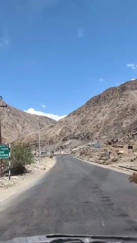 The video shows a moving vehicle's perspective as it travels on a paved road through a mountainous, arid landscape under a clear blue sky. The road curves gently to the left. On the left side of the road, there is a steep, rocky mountainside with sparse vegetation and power lines supported by wooden poles. A small settlement with buildings is visible in the distance. On the right side of the road, another steep, rocky mountainside rises. A guardrail with colorful panels, possibly depicting the Indian flag, runs along the edge of the road in the middle ground. The overall impression is of a journey through a remote, high-altitude region.