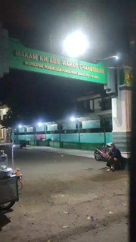 Here is a concise description of the video:

The video shows the entrance gate to the "MAKAM KH. ABD WAHAB CHASBULLAH," which translates to the tomb or burial site of a religious figure, Kh. Abd Wahab Chasbullah. The gate is green and white with the inscription in bold letters. The scene is at night, illuminated by streetlights.  A man in a white shirt and dark pants walks towards the gate. There is a food vendor's cart parked on the left side of the road near the gate.  Two people are sitting on the side of the road, and a scooter is parked nearby. The area around the gate looks somewhat deserted.