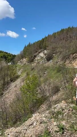 The video shows a hillside with a mix of trees and exposed rocky slopes. The trees are mostly deciduous with some evergreens, and many are just beginning to leaf out in bright green. The rocky slopes are covered with loose stones and some sparse vegetation. There is a small building visible on the hillside, and a tower on top of a distant hill. The sky is blue with some white clouds. The camera slowly pans up to show the landscape.